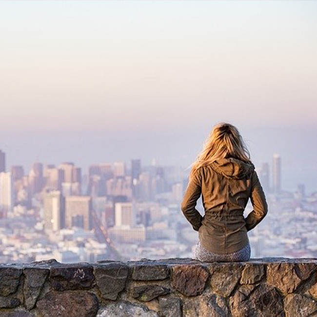 A woman sits with her back to the camera on a rock wall looking at a city in the distance