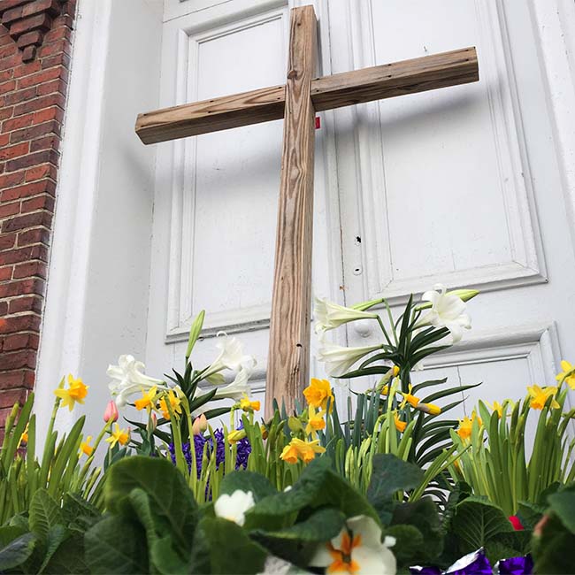 Close up of the front of the church with a large wooden cross resting against the door surrounded by flowers